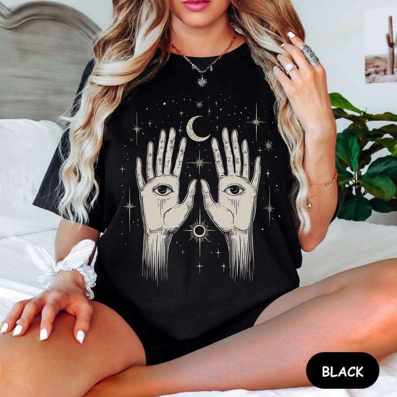 Może przedstawiać: Czarny T-shirt z grafiką dw&oacute;ch dłoni z oczami, rosnącego księżyca i gwiazd. Tekst "BLACK" jest widoczny w prawym dolnym rogu obrazu.