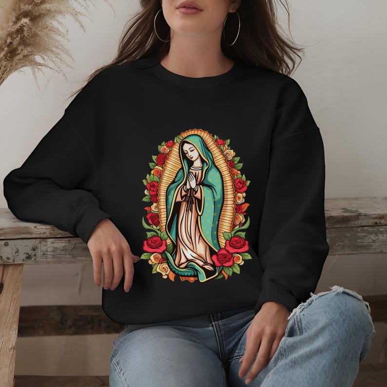 May include: Black sweatshirt featuring a colorful illustration of the Virgin Mary surrounded by roses. The artwork depicts a figure in a teal cloak and white dress, with hands clasped in prayer. The design is framed by a golden halo and red roses.