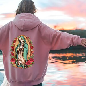 May include: A dusty rose hoodie featuring a graphic of the Virgin Mary surrounded by roses. The artwork is in a colorful, cartoon style. The hoodie has a drawstring hood and a kangaroo pocket.