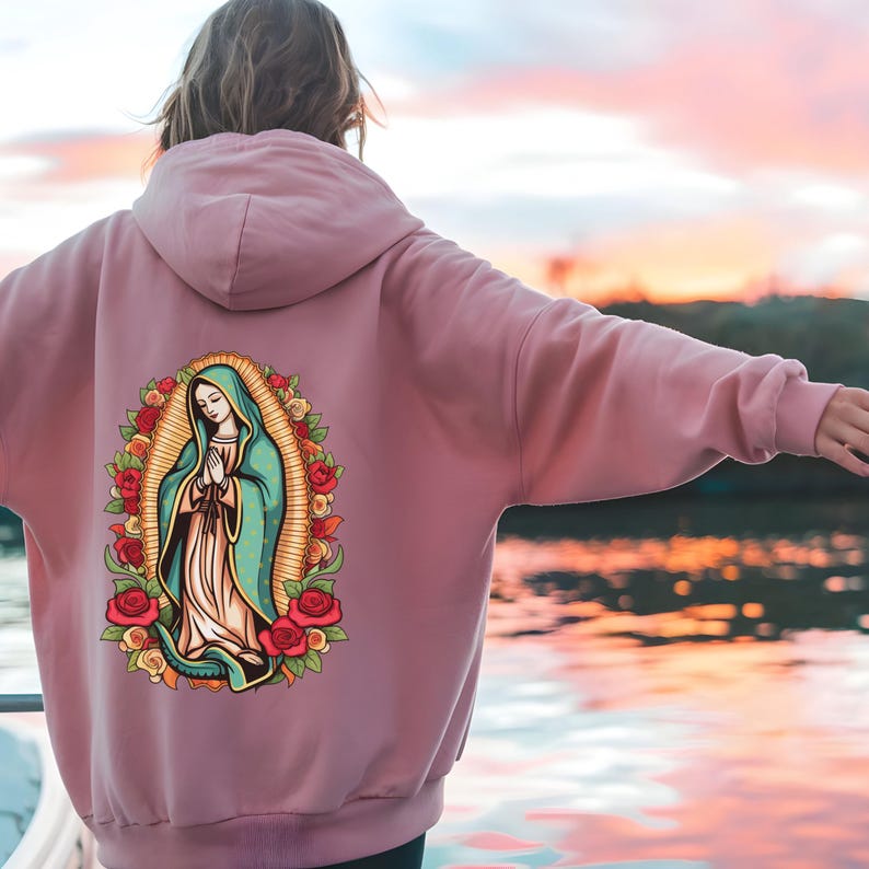 May include: A dusty rose hoodie featuring a graphic of the Virgin Mary surrounded by roses. The artwork is in a colorful, cartoon style. The hoodie has a drawstring hood and a kangaroo pocket.