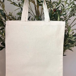 May include: A blank, off-white canvas tote bag with sturdy handles. The bag is rectangular and appears to be made of a durable fabric. The background features olive tree branches with green leaves and small olives.