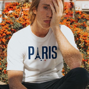 Può includere: Una persona giovane con le lentiggini indossa una maglietta bianca con una grafica blu della Torre Eiffel e la parola "PARIGI" stampata su di essa. La persona è seduta con la mano che le copre il viso.