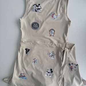 May include: A beige athletic dress featuring various embroidered Disney character patches. The patches include Mickey Mouse, Minnie Mouse, Dumbo, and others. The dress has a sleeveless top and a wrap-style skirt.