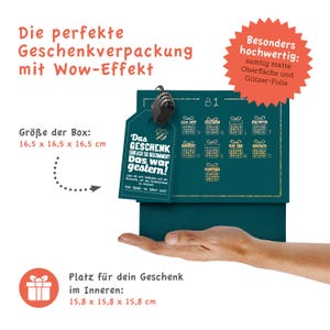 May include: A teal blue gift box with a gold glitter finish and a gold tag that says "Das Geschenk einfach so bekommen? Das war gestern!" The box is 16.5 cm by 16.5 cm by 16.5 cm. The box is sitting on a hand.