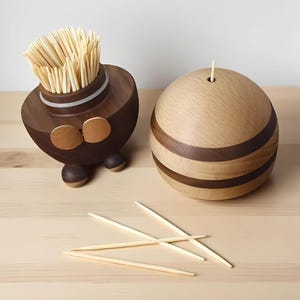 Könnte beinhalten: Zwei Zahnstocherhalter aus Holz. Einer ist dunkelbraun mit goldenen Akzenten und einem Stapel Zahnstocher. Der andere ist eine helle Holzkugel mit dunkelbraunen Bändern. Mehrere Zahnstocher sind auf der Oberfläche verstreut.