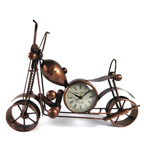 May include: A copper-colored metal motorcycle clock with a white face and black Roman numeral hour markers. The clock face reads "Village Clockmaker, Birmingham est. 1738".