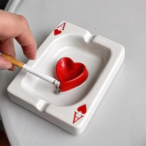 May include: A rectangular white ceramic ashtray shaped like a playing card, featuring the letter A and a red heart symbol in two corners. A raised, bright red heart-shaped basin sits in the center of the tray, holding a lit cigarette with ash.