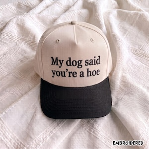 May include: A beige baseball cap with a black brim and black embroidered text on the front that reads, My dog said you're a hoe. The cap features a structured crown with ventilation eyelets and a button on top, showcasing a clean design.