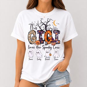 May include: A white t-shirt featuring a graphic with the text "This GIGI Loves Her Spooky Crew". The design includes large letters filled with Halloween patterns, a crescent moon, a tree, and four cartoon ghosts labeled James, Emily, Daniel, and Emma.
