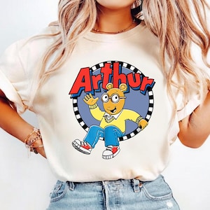May include: A cream-coloured short-sleeve t-shirt featuring a graphic of the cartoon character Arthur. The design shows Arthur waving inside a circular frame with a black and white checkered border, with the red text Arthur printed above him.