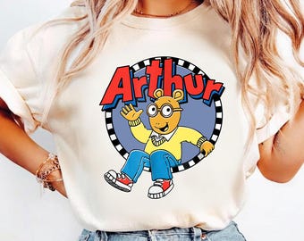 May include: A cream-coloured short-sleeve t-shirt featuring a graphic of the cartoon character Arthur. The design shows Arthur waving inside a circular frame with a black and white checkered border, with the red text Arthur printed above him.