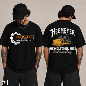 May include: A black t-shirt featuring a white gear graphic with the text Heemeyer Est. 2004 Demolition, Inc. on the front. The back displays a yellow bulldozer illustration with the text Heemeyer Est. 2004 Demolition, Inc. and the quote Sometimes reasonable men must do unreasonable things.