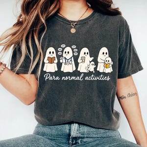 Peut inclure: Un t-shirt gris anthracite à manches courtes orné d'un motif représentant quatre fantômes blancs avec le texte "Para normal activities" en dessous. Les fantômes sont illustrés en train de lire, porter un stéthoscope et tenir un café.