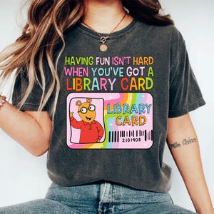 May include: Dark gray t-shirt with the text "HAVING FUN ISN'T HARD WHEN YOU'VE GOT A LIBRARY CARD." A pink library card graphic features a cartoon aardvark and rainbow accents.