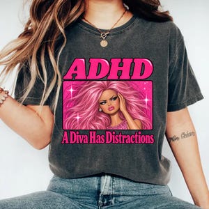 May include: A charcoal gray t-shirt featuring a graphic print with the text ADHD in bright pink letters above a rectangular frame. Inside the frame is an illustration of a character with long pink hair and the text A Diva Has Distractions below.