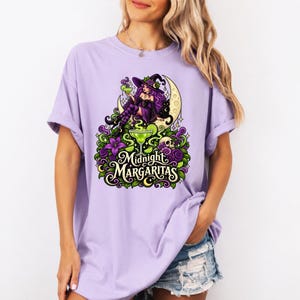 May include: A light purple t-shirt featuring a graphic print of a witch with purple hair sitting on a crescent moon, holding a margarita glass. Below, a larger margarita glass, a skull, and flowers are depicted with the text Midnight Margaritas.