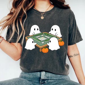 May include: Dark gray t-shirt featuring a playful Halloween design. The graphic shows four white ghost characters gathered around a green table playing mahjong, with two orange pumpkins. A fun, spooky design for the fall season.