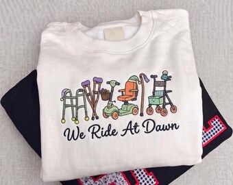 Könnte beinhalten: Ein weißes Sweatshirt mit Rundhalsausschnitt und einer Stickerei von Mobilitätshilfen wie einem Gehwagen, Krücken, einem Elektromobil und einem Rollator, die alle kleine Mützen tragen. Darunter ist der Text "We Ride At Dawn" in Schwarz gestickt.
