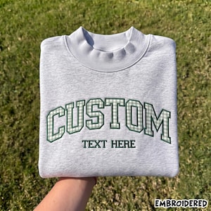 Può includere: Una felpa girocollo grigio chiaro con la parola CUSTOM ricamata in lettere a quadretti vichy verde scuro e bianco, con le parole TEXT HERE ricamate in verde scuro sotto. La felpa presenta un colletto a coste e maniche lunghe.