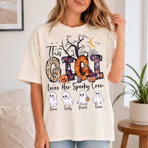 May include: A cream-colored t-shirt featuring a graphic with the text "This GIGI Loves Her Spooky Crew". The design includes a black tree, spiderwebs, a crescent moon, and four ghosts labeled with the names James, Emily, Daniel, and Emma.