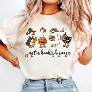 Pode incluir: Uma t-shirt de cor creme com um gráfico de quatro gansos vestidos com fantasias de Halloween, incluindo uma bruxa, uma abóbora, um fantasma e um esqueleto, todos segurando livros. Abaixo dos gansos, o texto diz "just a bookish goose".