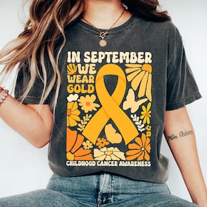 May include: A charcoal gray t-shirt featuring a large gold awareness ribbon in the center. The graphic includes flowers, butterflies, and the text "IN SEPTEMBER WE WEAR GOLD" and "CHILDHOOD CANCER AWARENESS" in a retro, rounded font style.
