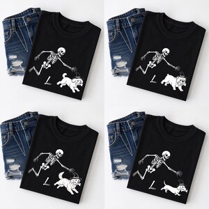 May include: Four black t-shirts each paired with blue denim shorts. Each shirt features a white graphic of a running skeleton reaching towards a dog wearing a witch hat and holding a bone. The dog breeds vary across the four shirt designs.