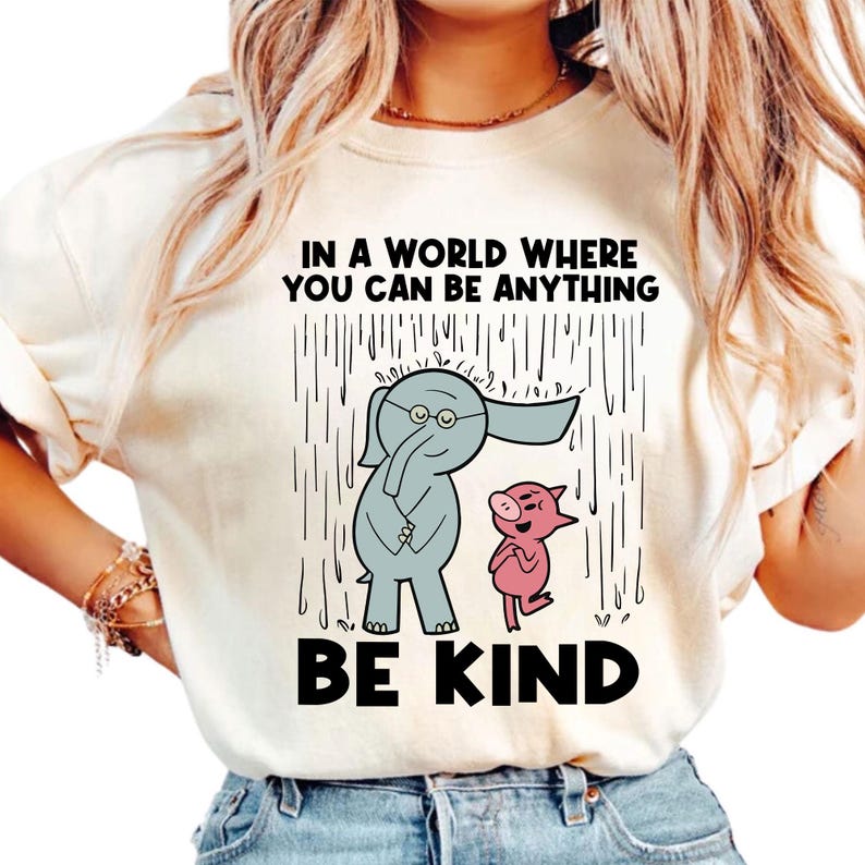 May include: A cream-coloured t-shirt featuring a graphic of a grey elephant and a pink pig standing in the rain. The text above reads IN A WORLD WHERE YOU CAN BE ANYTHING and the text below reads BE KIND. The design is centred on the chest.