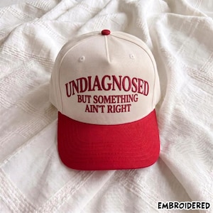 May include: A cream-colored baseball cap with a red brim and a red button on top. The front panel features red embroidered text that reads: UNDIAGNOSED BUT SOMETHING AIN'T RIGHT. The cap has a structured crown and a curved visor design.