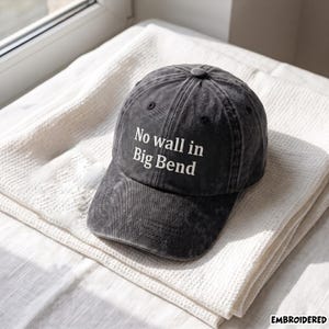 May include: A charcoal grey baseball cap with a distressed, washed-out finish. The front panel features white embroidered text that reads No wall in Big Bend. The cap has a structured crown, a curved brim, and a button at the top center.