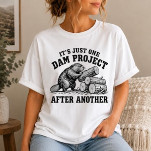 Puede incluir: Una camiseta blanca de manga corta con un gráfico negro de un castor apilando troncos. El texto sobre el castor dice IT'S JUST ONE DAM PROJECT y el texto debajo dice AFTER ANOTHER. El diseño está centrado en la parte frontal de la prenda.