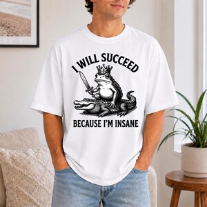 Op de afbeelding: Een wit T-shirt met korte mouwen en een zwarte afbeelding van een kikker met een kroon op die een zwaard vasthoudt terwijl hij op een alligator zit. De tekst erboven luidt I WILL SUCCEED en eronder staat BECAUSE I'M INSANE.