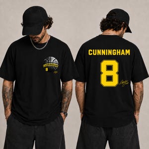May include: A black t-shirt featuring a basketball graphic on the left chest with the text CUNNINGHAM 8. The back displays the name CUNNINGHAM in yellow letters above a large yellow number 8 and a signature graphic in yellow ink.