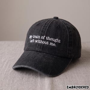 May include: A charcoal black baseball cap with a washed, distressed finish. The front panel features white embroidered text that reads, "My train of thought left without me." The cap has a curved brim and a button at the top center.