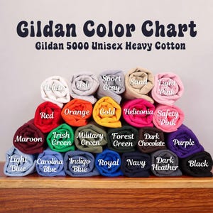 May include: A Gildan Color Chart for Gildan 5000 Unisex Heavy Cotton t-shirts, displaying a stacked arrangement of rolled fabric swatches in various colors, each labeled with its specific color name including White, Ash, Sport Gray, Sand, and Light Pink.