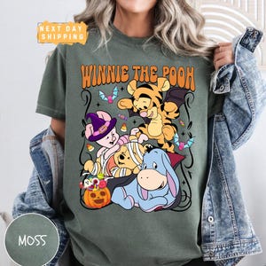 May include: A moss green t-shirt featuring a graphic of Winnie the Pooh characters dressed in Halloween costumes, including Piglet as a witch, Tigger as a bat, and Eeyore as a vampire, with the text WINNIE THE POOH and a jack-o'-lantern.