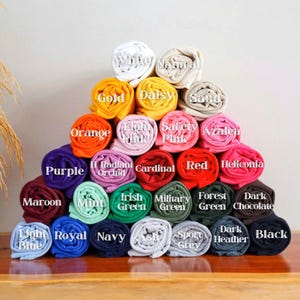 May include: A pyramid of rolled-up t-shirts in various colors, including white, gold, orange, purple, maroon, light blue, natural, daisy, light pink, radiant orchid, cardinal, red, heliconia, mint, irish green, military green, forest green, dark chocolate, sand, safety pink, azalea, royal, navy, ash, sport grey, dark heather, and black. Each shirt has a color label.