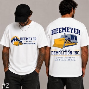 Peut inclure: Un t-shirt blanc avec un motif de bulldozer jaune et de montagnes sur le devant, accompagné du texte HEEMEYER DEMOLITION INC. EST. 2004. Le dos présente un plus grand graphique de bulldozer avec le même texte et la phrase Sometimes reasonable men must do unreasonable things.