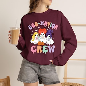 May include: A maroon long-sleeve sweatshirt featuring a graphic of three ghosts with different colored hair, two small pumpkins, and the text "BOO-HAVIOR CREW" in pastel colors. The design includes small white stars and bats surrounding the ghost figures.