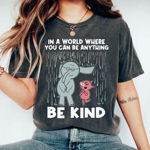 Pode incluir: Camiseta cinza escuro com o texto "IN A WORLD WHERE YOU CAN BE ANYTHING BE KIND." O gráfico apresenta um elefante cinza com óculos e um porco rosa, ambos sob um desenho de chuva.