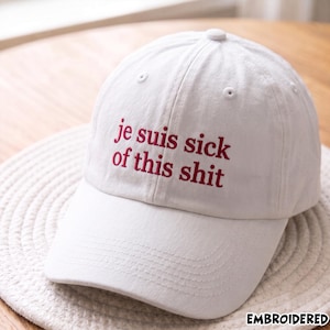 Pode incluir: Um boné de beisebol branco com o texto bordado "je suis sick of this shit" em fio vermelho escuro. O boné tem uma coroa estruturada, uma aba curva e ilhós costurados nos painéis superiores, proporcionando um design limpo e simples.