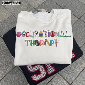 May include: A cream-colored crewneck sweatshirt featuring colorful embroidered letters that spell out OCCUPATIONAL THERAPY. Each letter is creatively formed by various therapy-related icons, including a yoga ball, a wheelchair, and a brain.