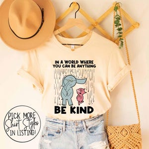 May include: A cream-coloured t-shirt featuring a graphic of a blue elephant and a pink pig standing in the rain. The text above reads IN A WORLD WHERE YOU CAN BE ANYTHING and the text below reads BE KIND. The shirt is displayed on a wooden hanger.