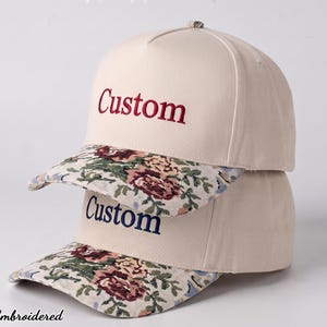 May include: Two stacked baseball caps in a light cream color. The top cap features the word Custom embroidered in red, while the bottom cap features the word Custom in navy blue. Both caps have a floral patterned brim with roses and green leaves.
