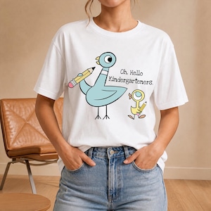 Può includere: Una t-shirt bianca a maniche corte con la grafica di un piccione azzurro e un piccolo anatroccolo giallo, entrambi con in mano matite gialle. Il testo "Oh, Hello Kindergarteners." è stampato in nero accanto al piccione.