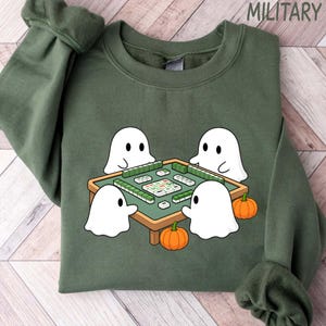 May include: Green sweatshirt with a graphic of four white cartoon ghosts playing mahjong around a green table. Two orange pumpkins are at the bottom. The word "MILITARY" is printed at the top.