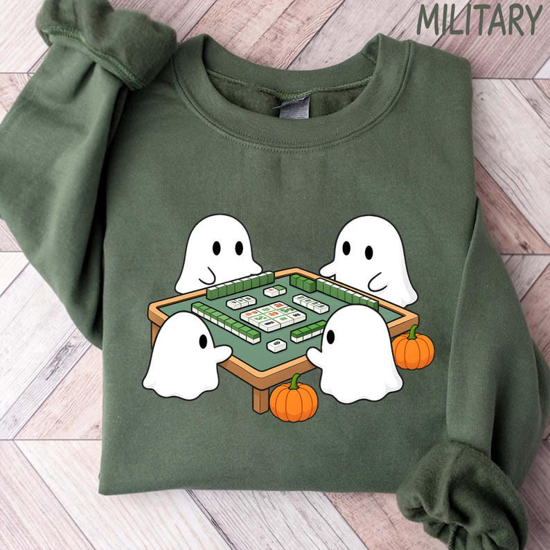 May include: Green sweatshirt with a graphic of four white cartoon ghosts playing mahjong around a green table. Two orange pumpkins are at the bottom. The word "MILITARY" is printed at the top.