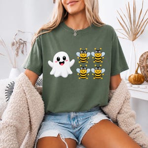 May include: An olive green short-sleeve t-shirt featuring a graphic print of a white cartoon ghost positioned to the left of four small, smiling yellow and black striped bees arranged in a two-by-two grid pattern on the front of the garment.