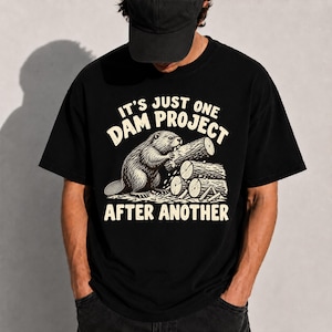 Könnte beinhalten: Ein schwarzes T-Shirt mit kurzen Ärmeln und einer Grafik eines Bibers, der an einem Holzstamm nagt. Der Text IT'S JUST ONE DAM PROJECT AFTER ANOTHER ist in Cremefarben gedruckt. Das Motiv zeigt zudem Holzstämme und Holzspäne am Boden.