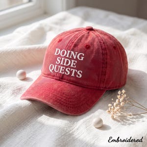 Könnte beinhalten: Eine rote Baseballkappe mit Used-Look und dem weiß gestickten Text DOING SIDE QUESTS auf der Vorderseite. Die Kappe hat einen gebogenen Schirm, einen Knopf oben und genähte Ösen. Das Wort Embroidered ist in der Ecke aufgedruckt.